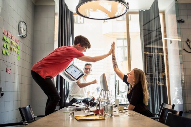 2 collega's geven elkaar een high-five. Een van hun staat op een stoel. Een derde collega glimlacht achterin de ruimte.